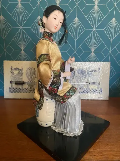 Poupée Geisha Japonaise. Début XXème, tête et main en porcelaine, ainsi que le pot lui servant de siège. Sur socle en bois laqué noir. Très élégante et en bon état.    Japanese Geisha doll. Early 20th century, porcelain head and hand, as well as the pot serving as a seat. On a black lacquered wooden base. Very elegant and in good condition.